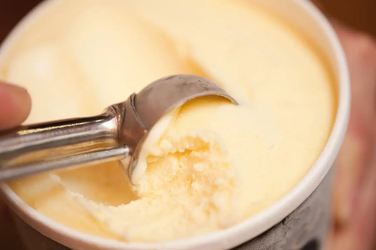 A hand scooping creamy vanilla ice cream from a carton with an ice cream scoop.